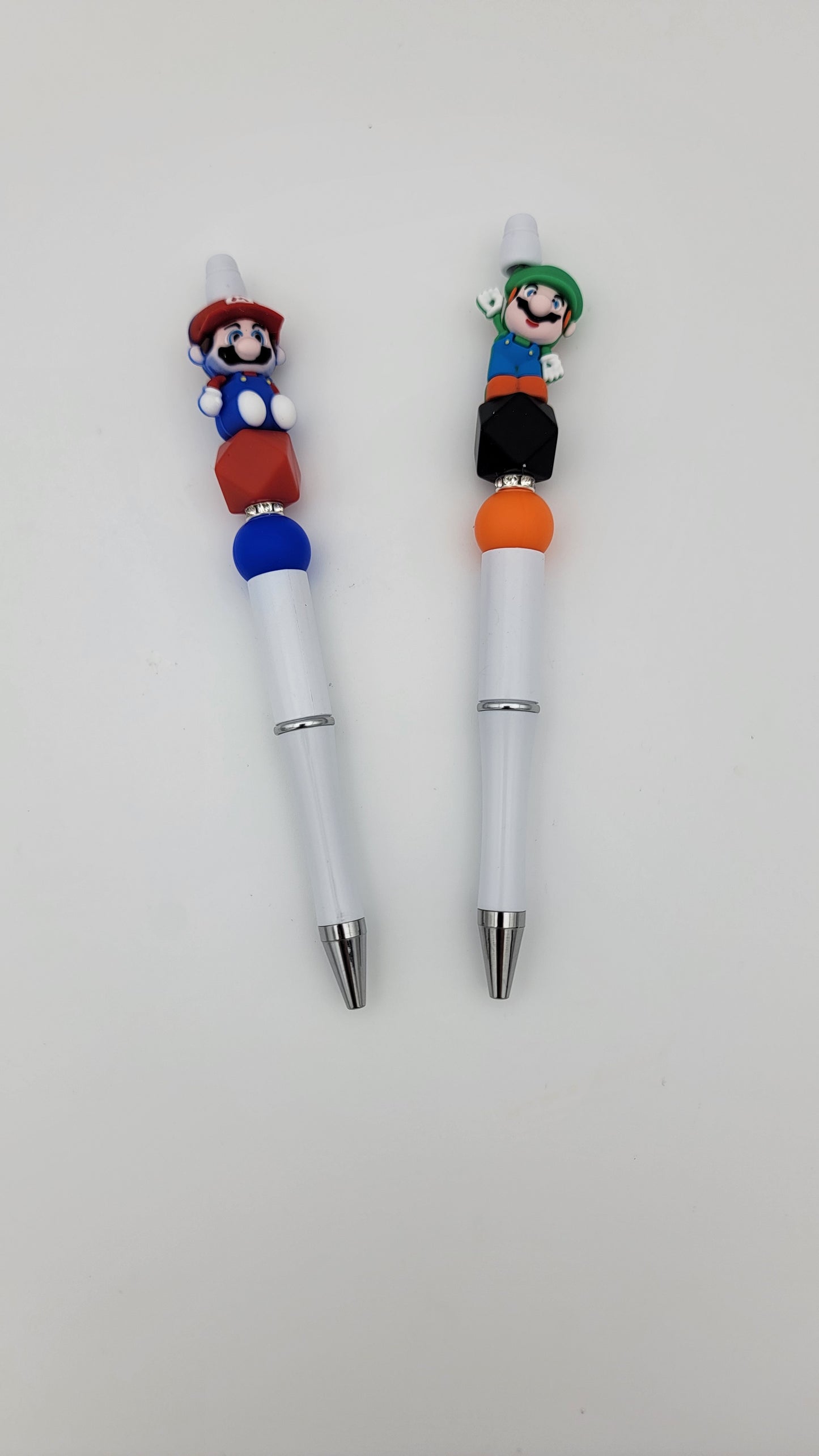 Two pens with decorative toppers of animated characters in red and green hats, featuring colorful beads, set against a plain white background. Product: Beaded Pens-2 in 1 bag.