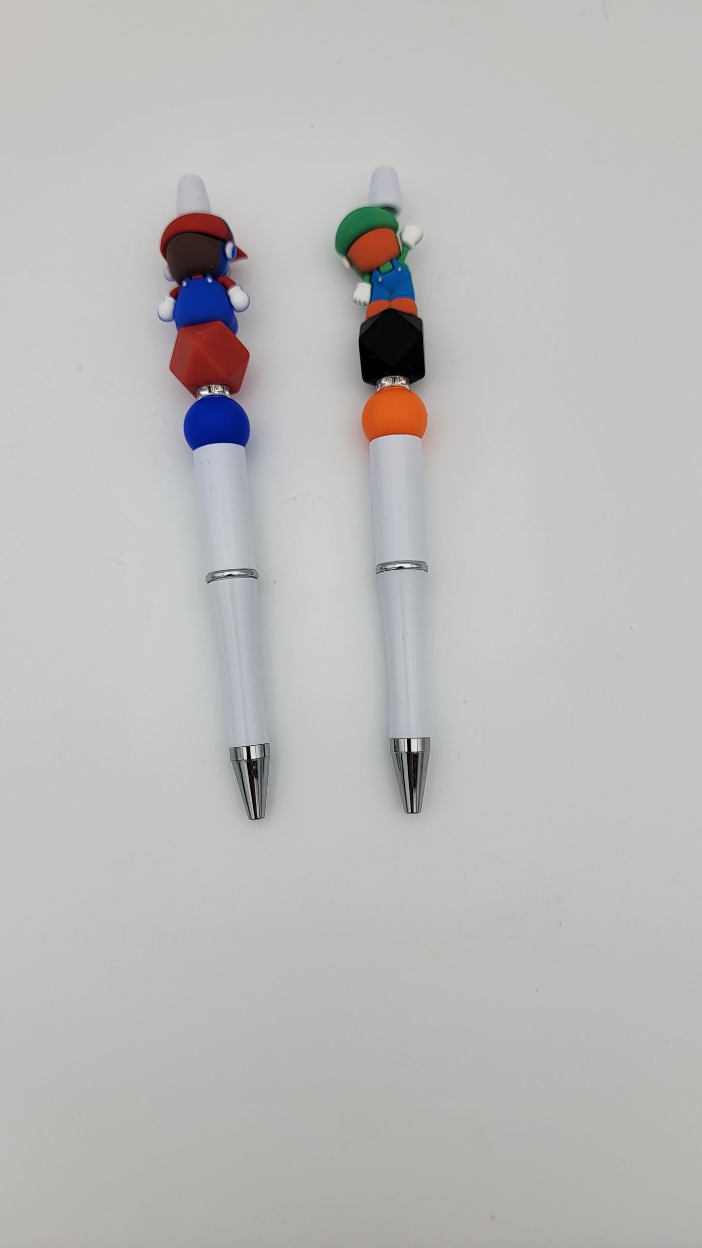 The Beaded Pens-2 in 1 bag includes two white pens, each with a metallic tip and decorative topper. One pen features blue and red shapes, while the other has green, orange, and black shapes.