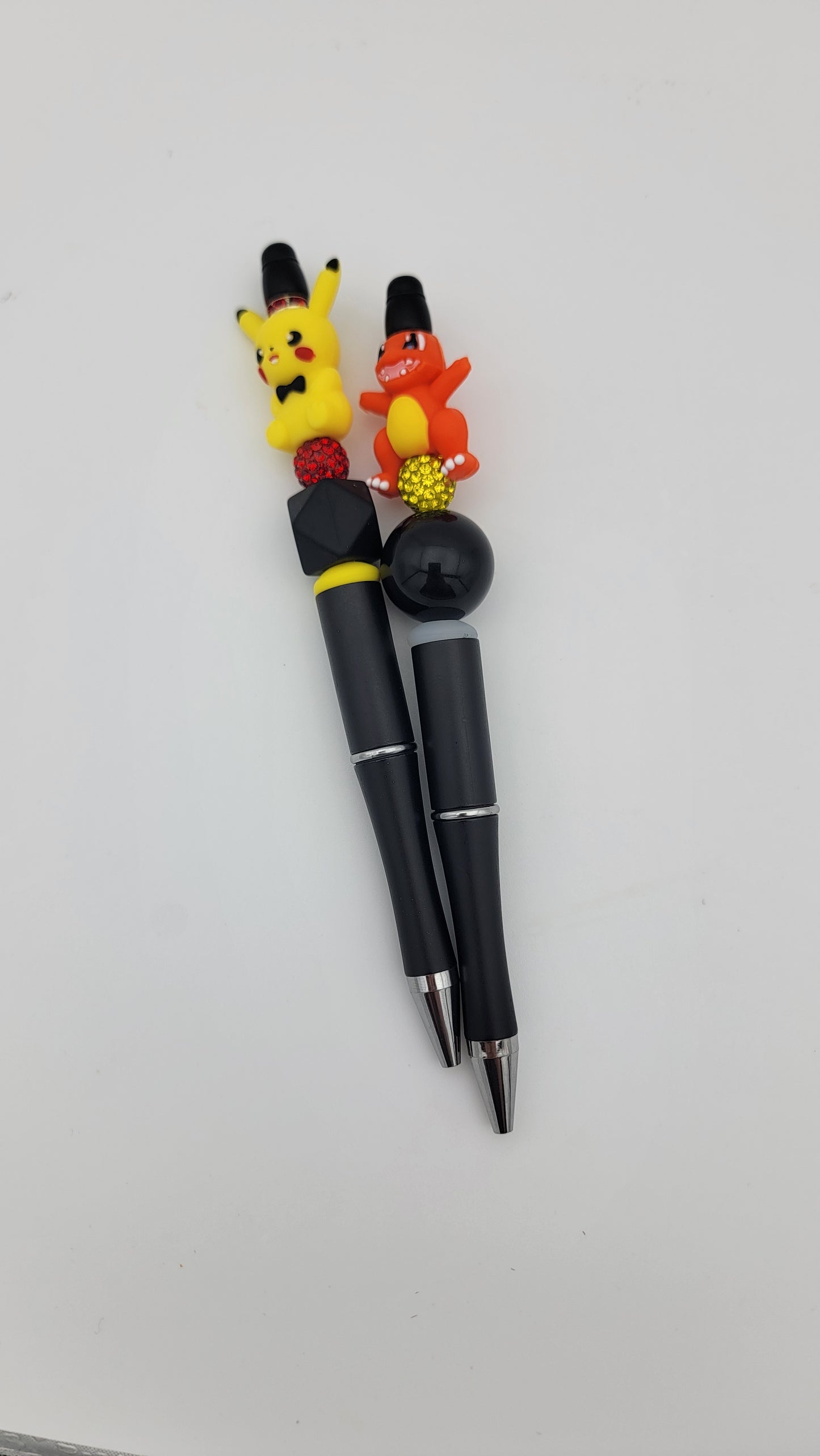 Two black Beaded Pens, each topped with a colorful toy depicting popular cartoon creatures, one yellow and the other orange, are set against a white background.