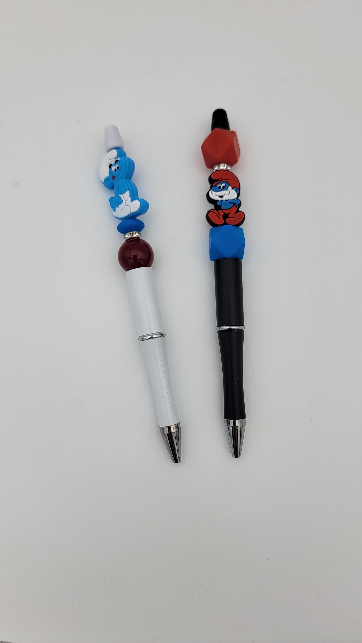 Two Beaded Pens with decorative Smurf figures—the left one is white and the right one is black—are shown against a plain white background.
