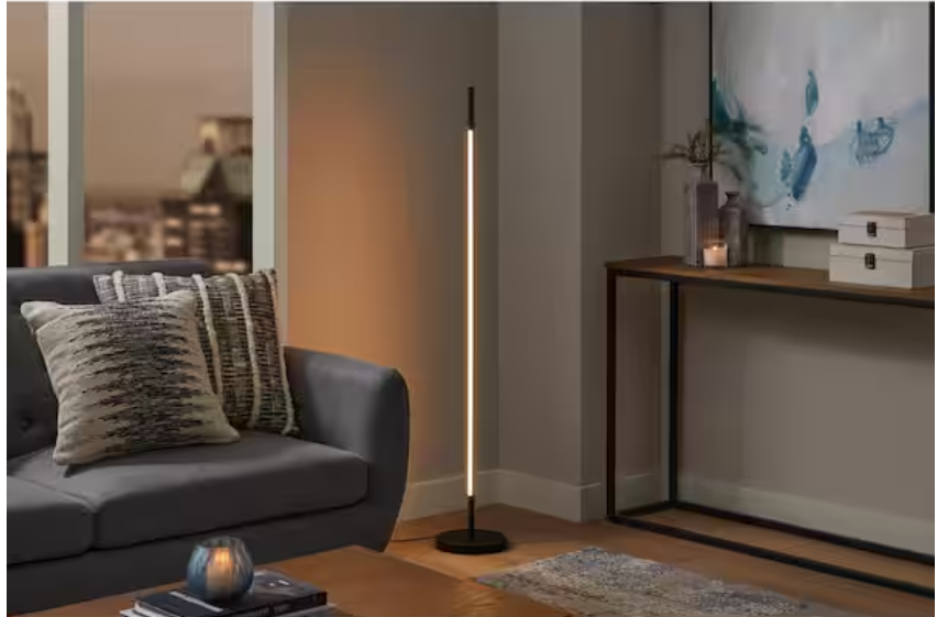 Modern living room with a gray sofa, striped cushions, a Hampton Bay 50" Calero Black Floor Lamp, wooden console table, abstract wall art, and a cityscape view in the background.