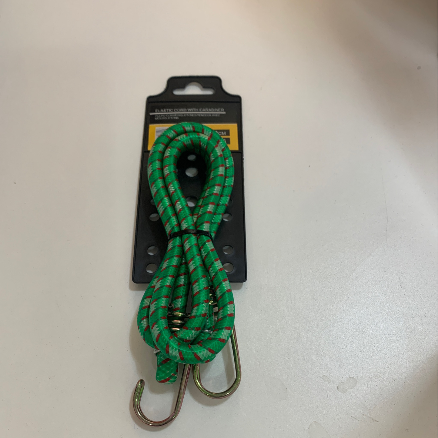 A green bungee cord with red accents and metal hooks is placed on black packaging featuring a yellow and black label, resting on a white surface. Product: Bungie cord w carabiner.