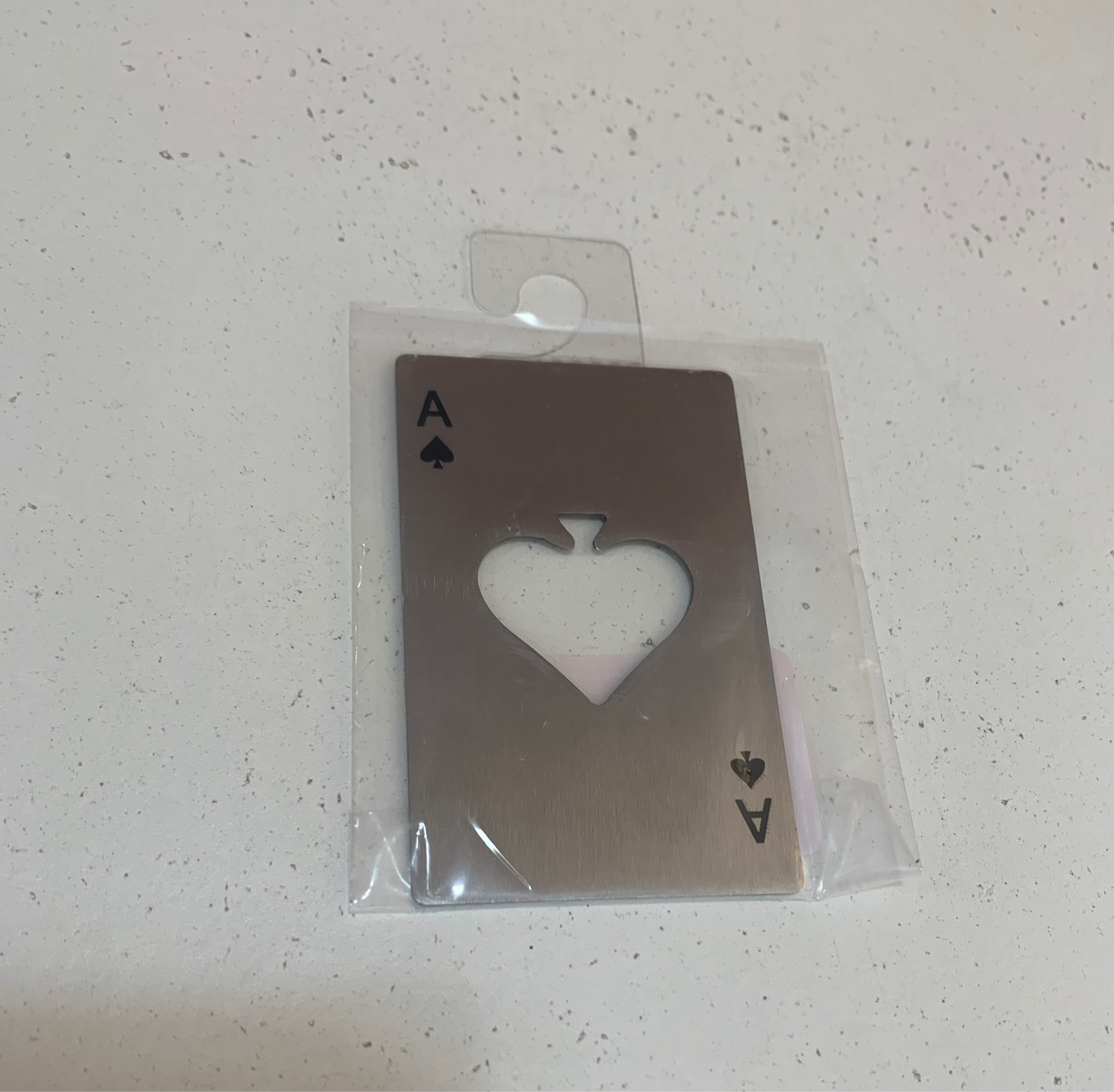 An Ace card bottle opener, featuring a heart-shaped cutout in the center, is encased in a clear plastic sleeve and rests on a speckled surface.