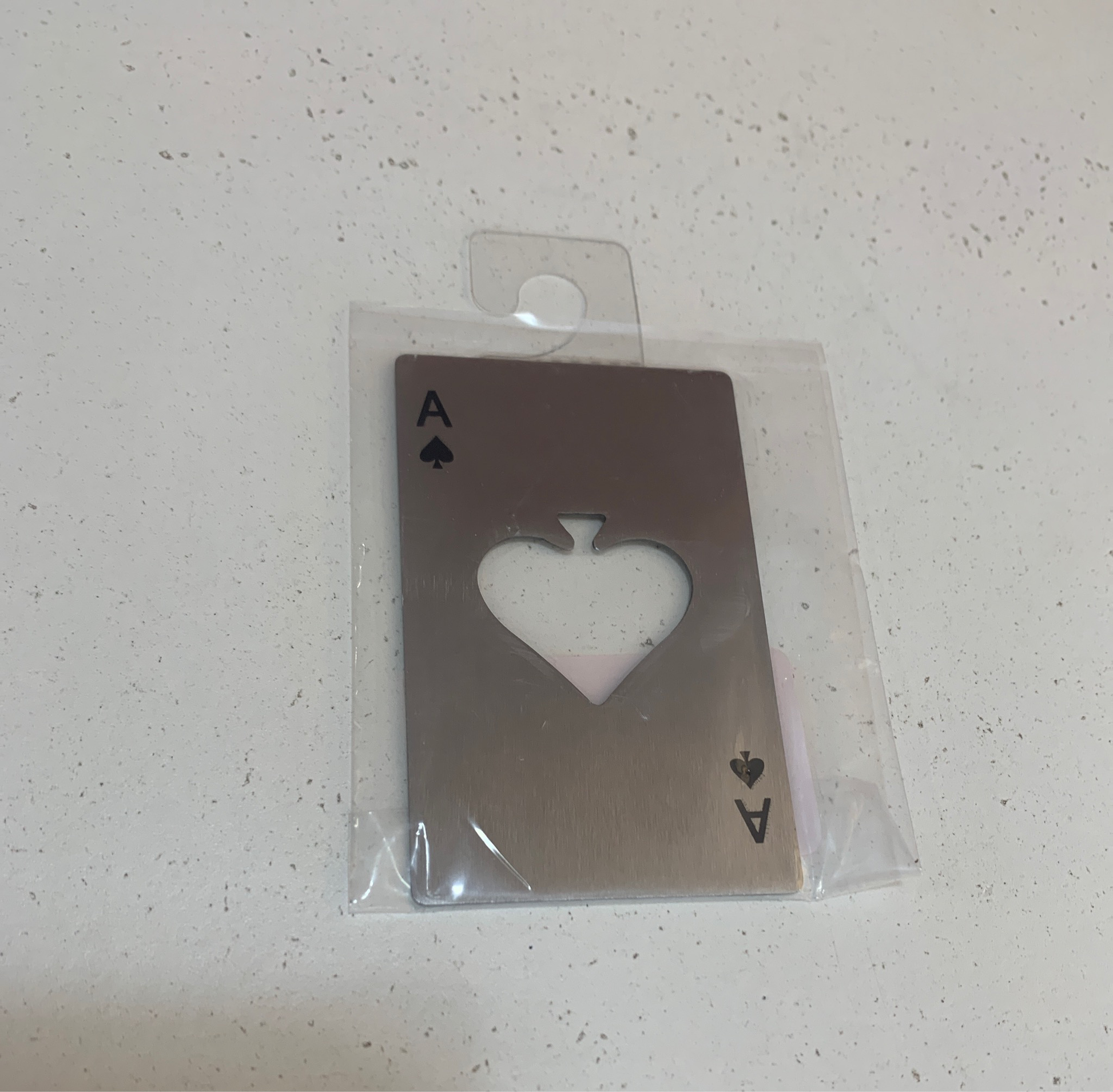 An Ace card bottle opener, featuring a heart-shaped cutout in the center, is encased in a clear plastic sleeve and rests on a speckled surface.