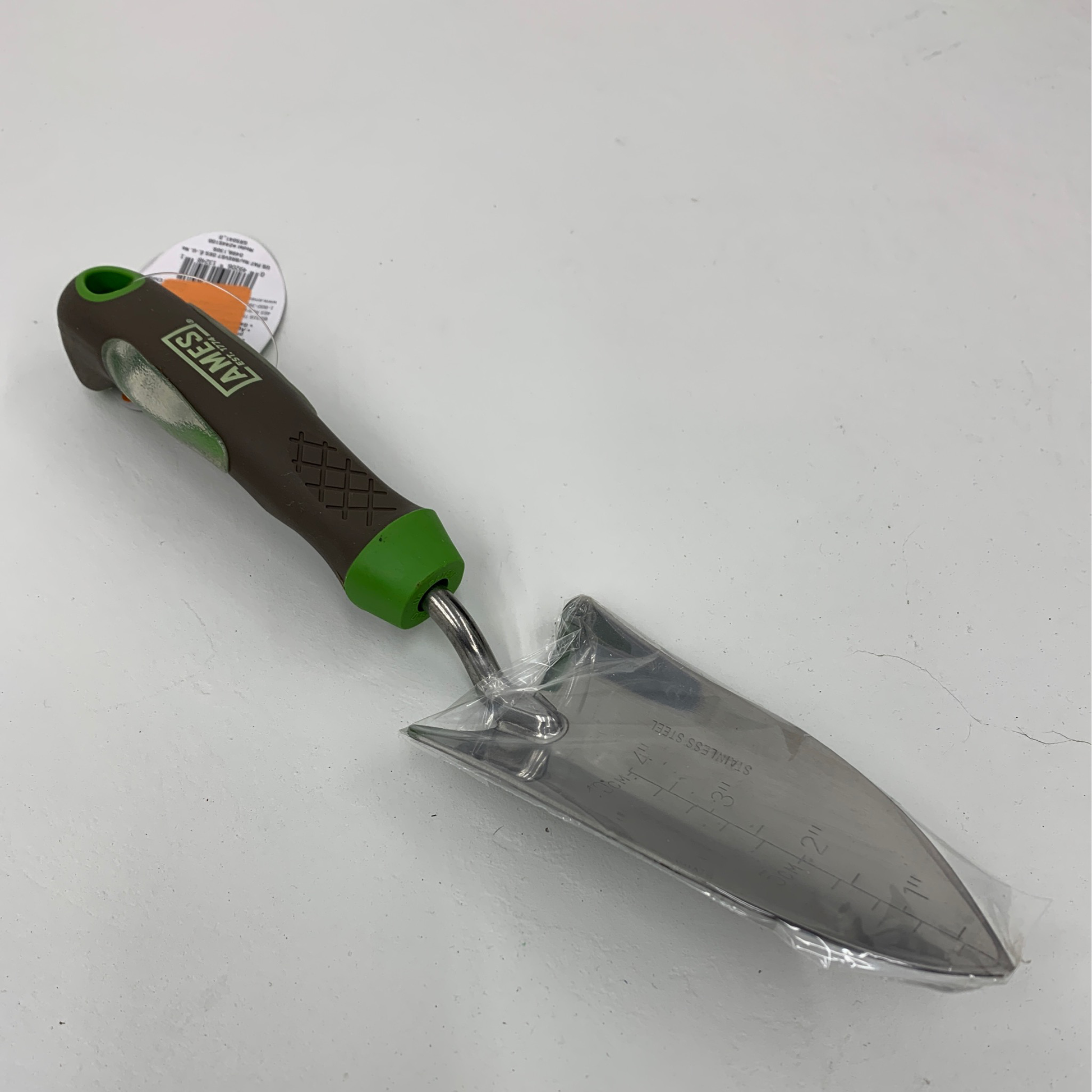 The Ames ergo gel grip transplanter, with a green and brown handle and measurement markings on its silver blade, is displayed on a light surface.