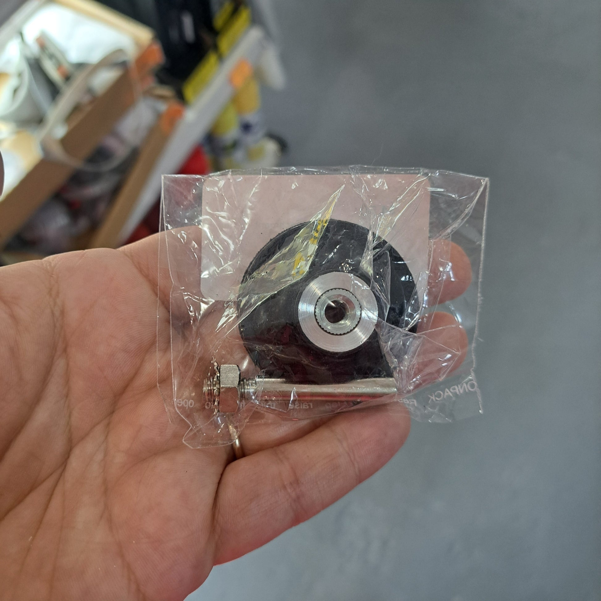 A hand holding a scouring pad drill attachment wrapped in plastic, with accompanying screw and nut, set against the backdrop of a store aisle lined with shelves.
